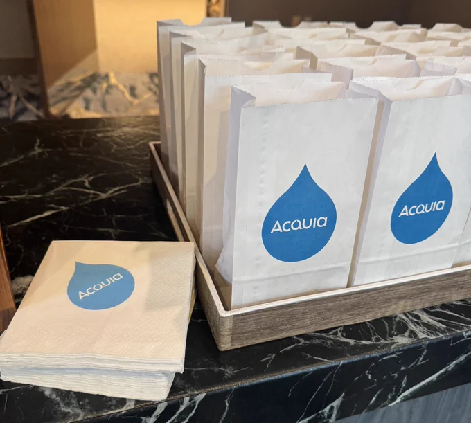 White paper bags with a blue water drop logo labeled 'Acquia' neatly arranged in a wooden tray on a black marble surface.