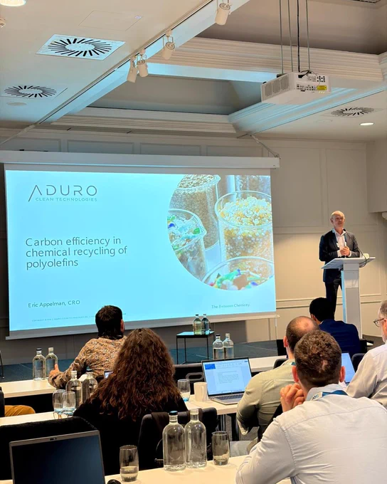Eric Appelman standing at a podium in front of an attentive audience. A powerpoint slide is shown on a projector screen. The title reads “Carbon efficiency in chemical recycling of polyolefins.