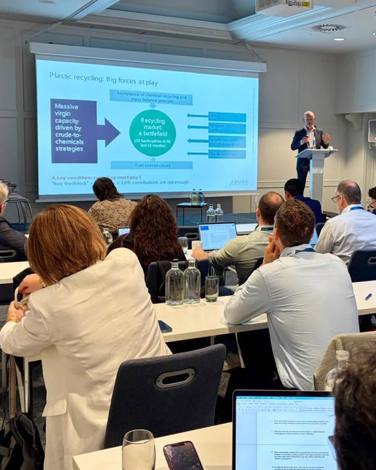 Eric speaking at a podium in front of an attentive audience. The powerpoint shows a flow chart explaining the forces at play in plastic recycling. 