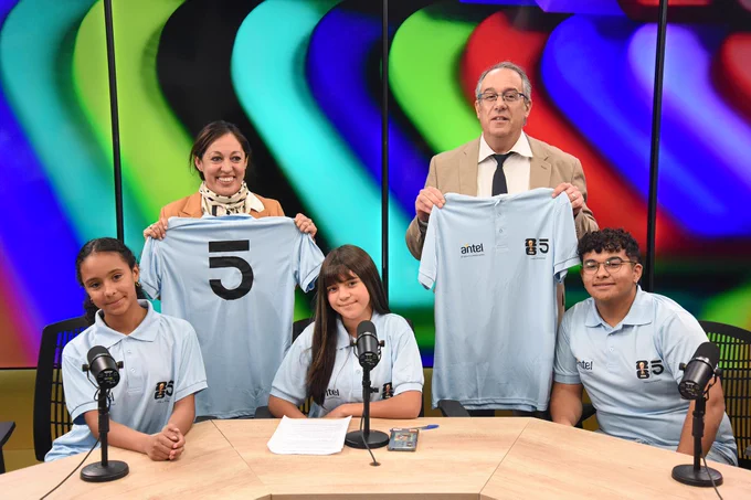 Adolescentes sentadas (dos) y sentado en mesa de estudio; visten camisetas celestes. De pie, ministra Fernanda Cardona y ministro José Carlos Mahía sostienen camisetas de Uruguay. Detrás, pantalla con colores.
