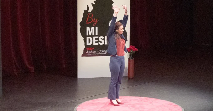 Dr. Elizabeth Doxtader  on a red circular carpet on stage with arms raised during a TEDx talk at Jackson College.