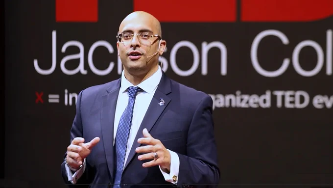 Speaker Kamal Ghazi delivering a TED Talk on stage with a large screen displaying text behind him.