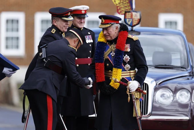 A group of uniformed individuals are standing in front of a parked car with a regal emblem. They are engaged in a formal, cordial interaction, with one person bowing slightly while shaking hands. The individuals are adorned with various ceremonial decorations. A building with white-trimmed windows is visible in the background.