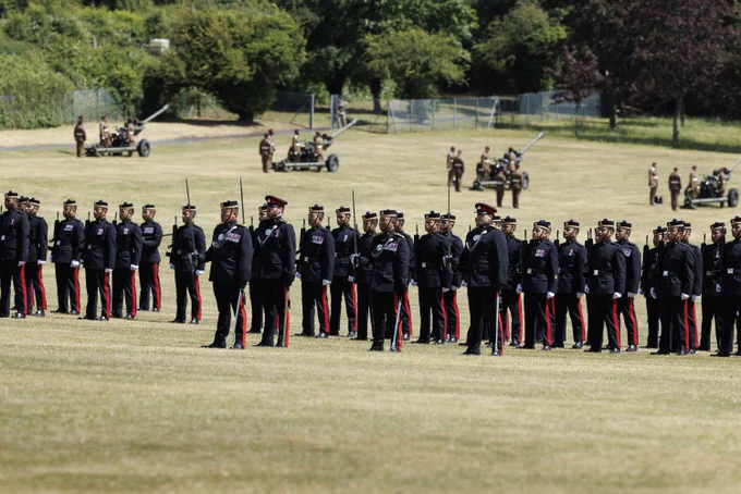 A group of soldiers in formal uniforms stands in formation on a grassy field. In the background, several military personnel are positioned near artillery pieces. Trees and fences are visible beyond the field.