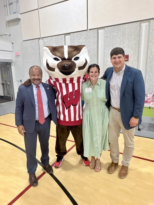 A group of three people (Interim Chancellor Wilcots, Jeannie Cullen Schultz, and George Cullen) stands indoors on a polished wooden floor, posing with Bucky Badger. Bucky is dressed in a red and white striped shirt with a red "W.