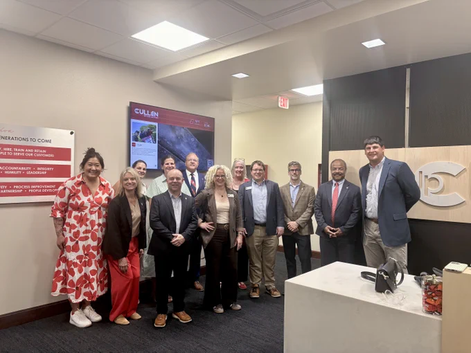 A group of people (including Interim Chancellor Wilcots, UW leaders, Cullen staff and Wisconsin Legislators) stand together in an office setting, posing for a photo. They are dressed in business attire. Behind them is a board with information and a digital display featuring "JP Cullen." The room has neutral-colored walls.