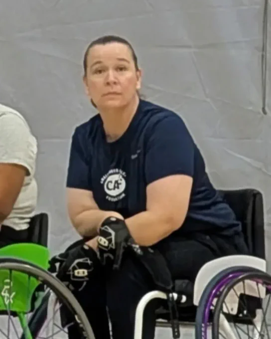 Person in a wheelchair wearing a navy shirt and gloves, looking forward, with a gray background.