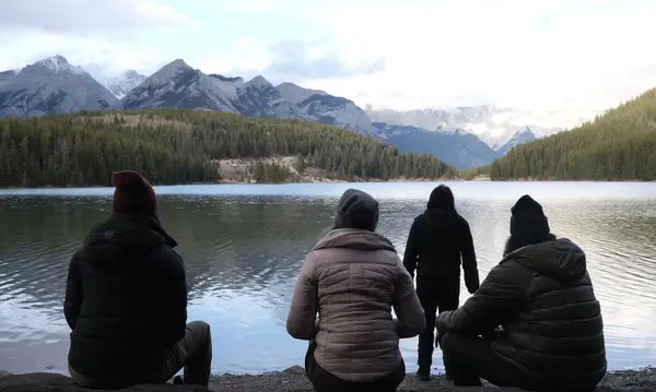 A still from the film Meadowlarks. Four First Nations people - a man and three women - stand on the shore of a shimmering silver lake surrounded by forest, with mist-shrouded mountains beyond it.
