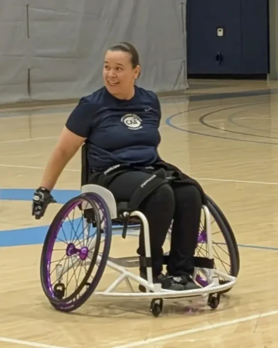 Person in a sports wheelchair on a gym floor, smiling and wearing a navy shirt with a logo.