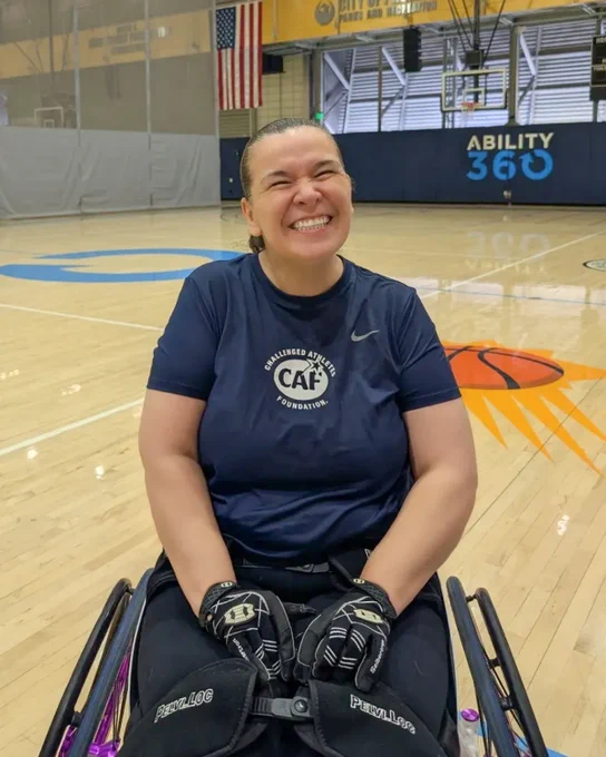 Smiling woman in a wheelchair on a basketball court, wearing a "Challenged Athletes Foundation" shirt.