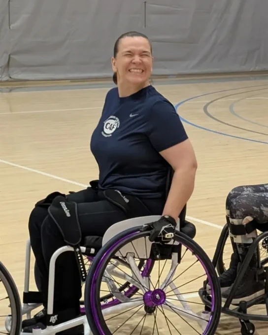 Smiling woman in a sports wheelchair on a gym court, wearing a navy shirt and gloves.