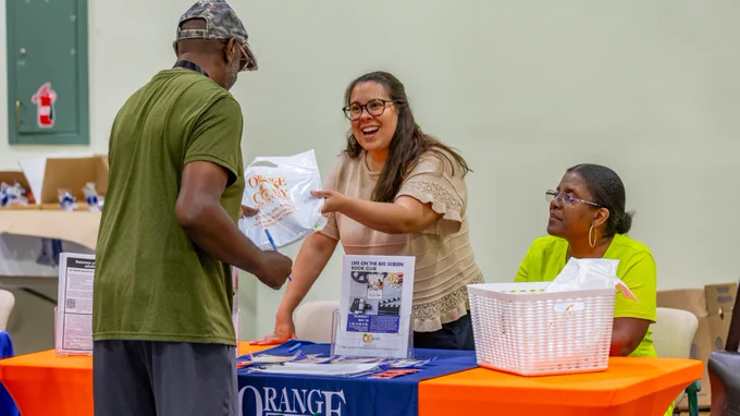 A person hands a bag to another person across a table covered with an "Orange County Government" branded tablecloth. A third person sits and observes. The table holds informational materials on Office on Aging with a white basket filled with items.