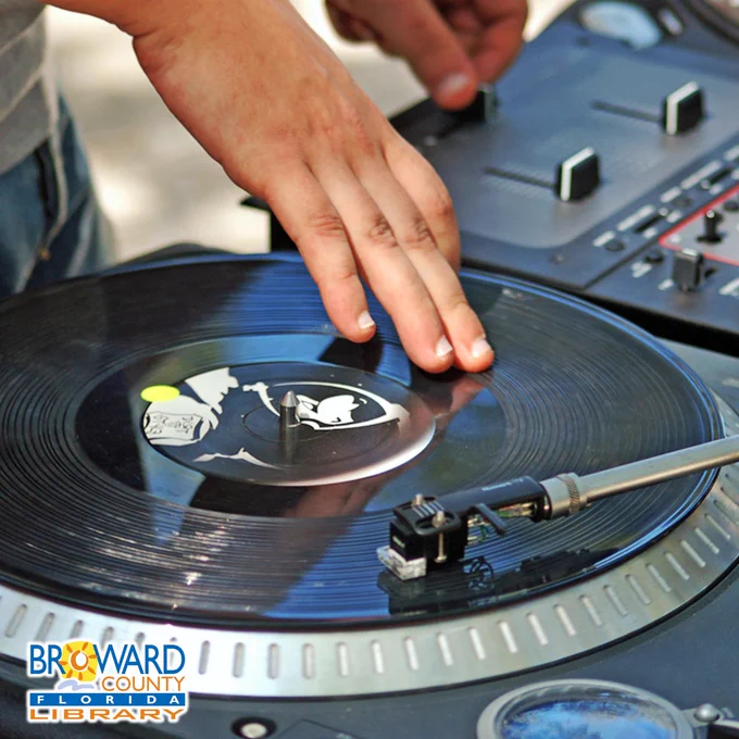 Close-up of a person's hands operating DJ equipment. One hand rests on a spinning black vinyl record on a turntable with the needle down, while the other hand adjusts faders on a mixing board in the background. The Broward County Florida Library logo is in the bottom left corner.