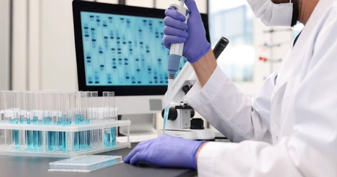 A person in a lab coat and gloves uses a pipette at a laboratory workstation. There are test tubes with blue liquid on a rack, and a computer screen displaying a DNA sequence in the background.