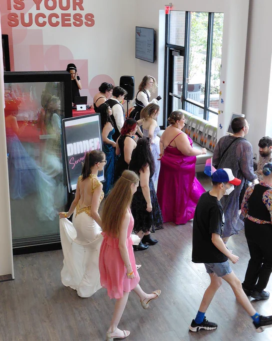 A group of people in formal attire walk through a room with large windows. Some hold their dresses as they proceed in a line. There's a sign with the words "OUR GOAL IS YOUR SUCCESS" near the entrance.