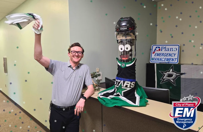 A smiling man with glasses and a gray polo shirt waves a white cloth in his hand, standing next to a tall object. The object, decorated to resemble a cartoon character, wears a hockey helmet and a Dallas Stars jersey. A sign in the background reads "Plano EM Emergency Management," with a logo. 