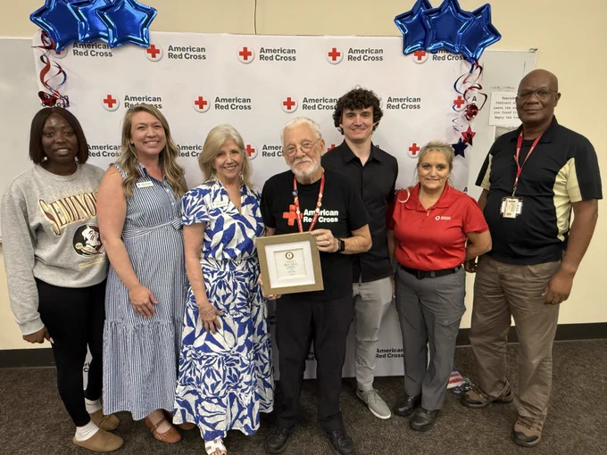 A group of seven people stands together, smiling, in front of an American Red Cross backdrop with balloons. The person in the center holds an award plaque. The background includes multiple American Red Cross logos.