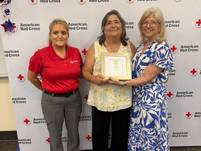 Three individuals are standing in front of an "American Red Cross" backdrop. The person in the center is holding a framed certificate. The individual on the left is wearing a red shirt with the Red Cross logo, while the person on the right is in a blue and white patterned outfit.