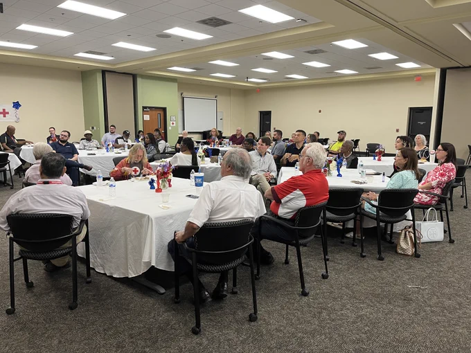 A group of people seated around tables in a conference room, attentively listening to a speaker. The room is well-lit, and the tables are decorated with red, white, and blue centerpieces. The audience appears engaged, with some taking notes.