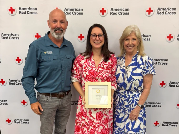 Three people stand smiling in front of an American Red Cross backdrop. The person in the middle holds a framed certificate.