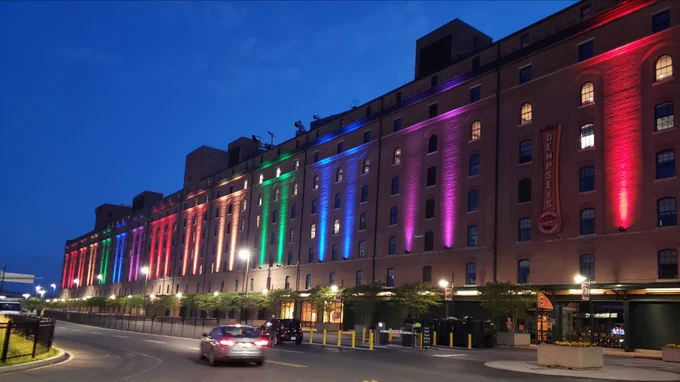 The Warehouse at Camden Yards is illuminated with colorful vertical lights in rainbow hues at dusk and the sky is a deep blue. 