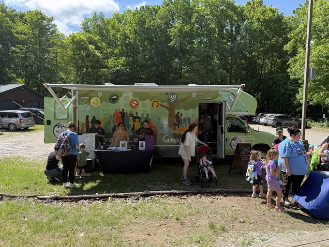 A mobile library truck is parked outdoors on a sunny day, with groups of people gathered around tables displaying books and brochures. The truck features colorful graphics of diverse individuals and social media icons. Families and individuals appear to be engaging with the materials and enjoying the event. Trees and a parking area filled with vehicles are visible in the background.
