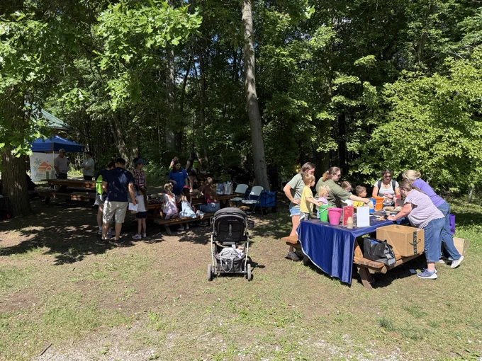 A group of people is gathered outdoors under trees, some sitting at picnic tables and others standing near tables with items. A stroller is in the foreground, and there are chairs set up nearby. It appears to be a community event or gathering.
