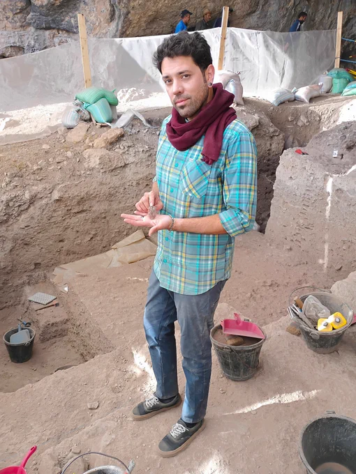 Dr. Ismael wearing a plaid shirt and scarf stands in an archaeological dig site surrounded by excavation tools and dirt.