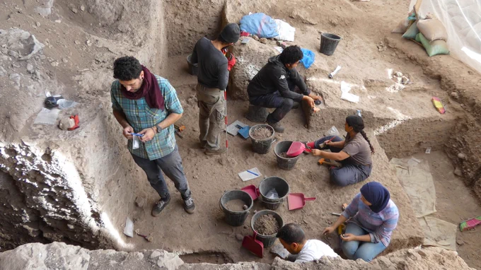 Archaeologists, including Dr. Ismael, carefully excavate and document artifacts at a dig site with tools and buckets scattered around.