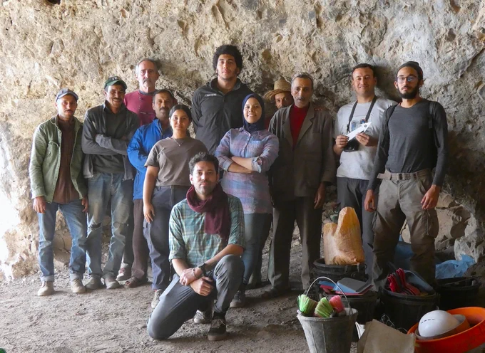 Group of ten people posing inside a rocky cave with various outdoor gear and supplies on the ground nearby in Morocco.