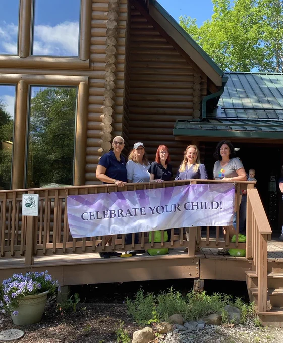 Five people stand on a wooden ramp in front of a log building, holding a banner that reads, "Celebrate Your Child!" The setting is outdoors with greenery and a clear sky.
