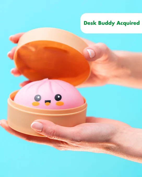 Hands holding an open round container with a smiling pink dumpling toy inside against a blue background. Text says "Desk Buddy Acquired."