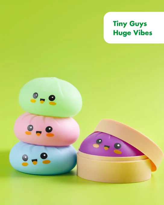 Three pastel-colored squishy toys with smiling faces stacked beside a purple squishy in a round beige container against a green background. Text says "Tiny Guys Huge Vibes"