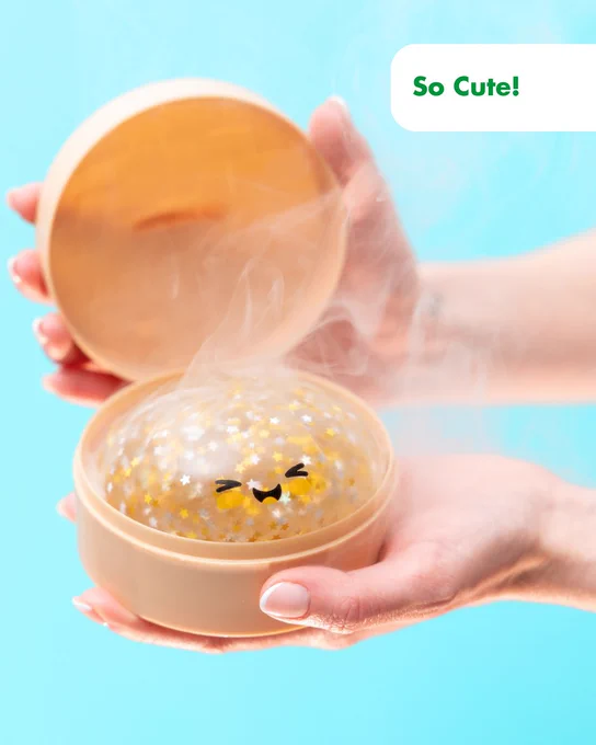 Hands holding an open round container with a steaming, smiling gel-like ball decorated with golden stars inside. Text says "So cute