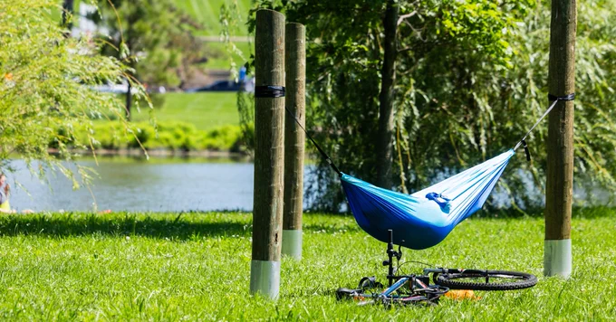 A person inside a blue hammock is hanging from human-made poles as their bike lays on it's side in the grass while full-leaf trees nearly obscure a waterway behind them. 