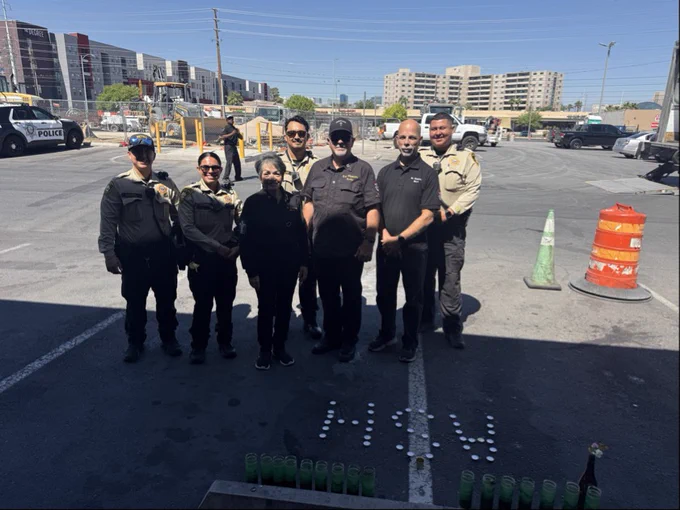 Group of uniformed LVMPD officers and chaplains standing together in a parking lot in front of candles and the name Ash spelled out on the ground. There are buildings and police vehicles in the background.