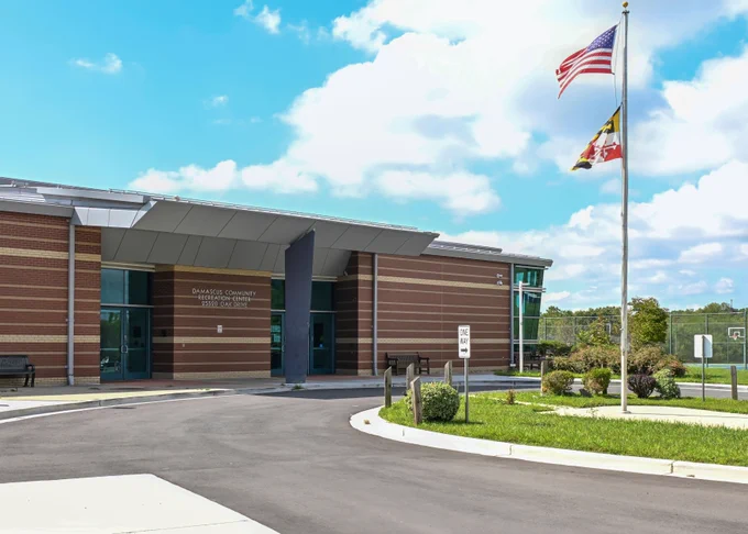 Modern community center building with striped brick exterior under a partly cloudy sky and two flags flying on a tall flagpole.