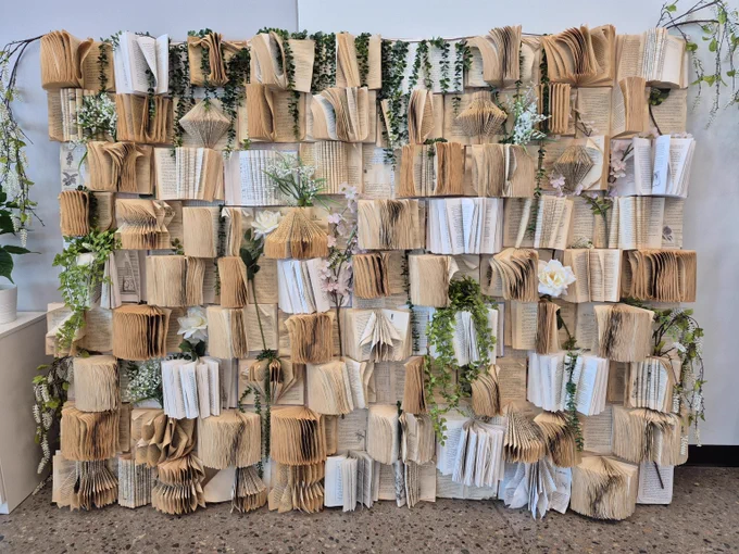 Wall art installation made of numerous open books with folded pages, interspersed with green vines and white flowers for a natural aesthetic.
