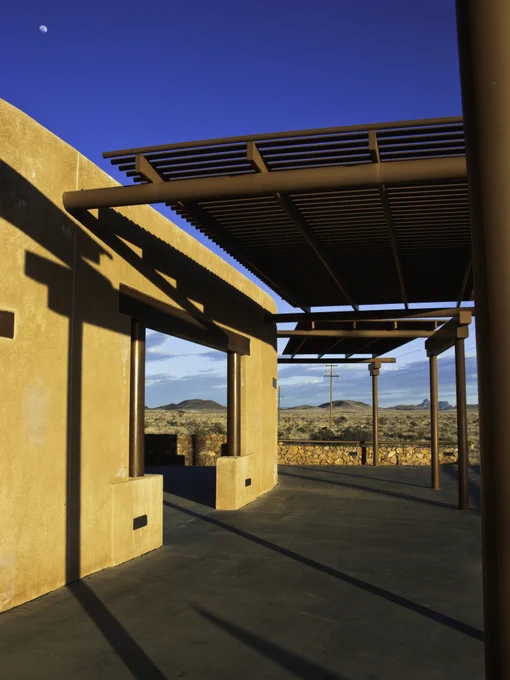 Modern adobe-style building with shaded patio overlooking a desert landscape under a clear blue sky.