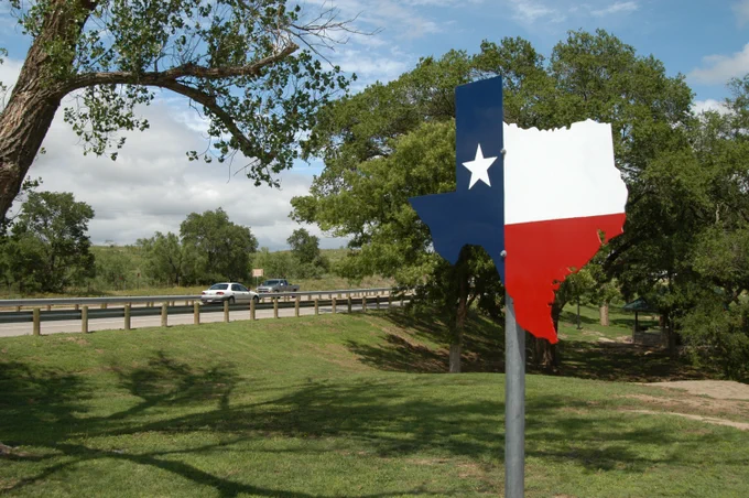 Large metal Texas state silhouette painted with the Lone Star flag colors standing on a grassy roadside under a blue sky.