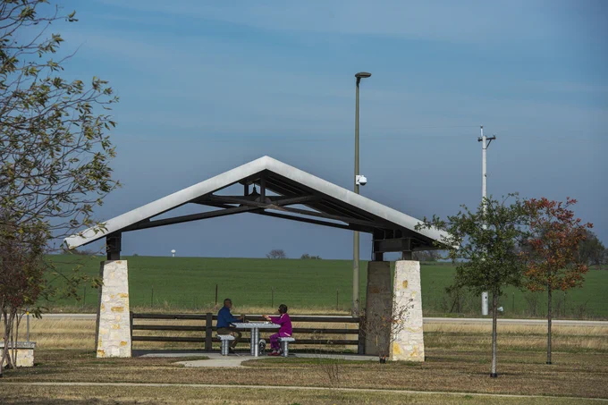 Two people sitting and talking at a picnic table under a small pavilion in a grassy park area on a clear day.