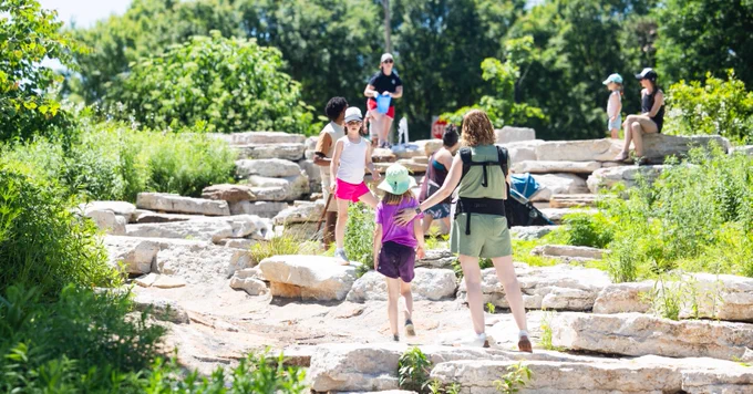Flaked by green foliage, a child wearing a bright purple shirt is centered among others while exploring the rocky landscape of a human-made spring as full-leaf trees fill the background.