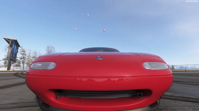 Un coche deportivo rojo, está situado en una pista con un cielo azul claro. Varios globos aerostáticos flotan en el fondo.