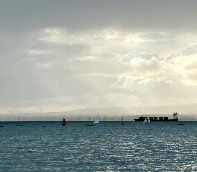 A seascape. A large tanker passes from right to left behind a group of small sailing boats. The water is choppy, and a dark turquoise. The hills on the opposite side are mistily shrouded by the clouds, through which fingers of light faintly shine down towards the tanker.