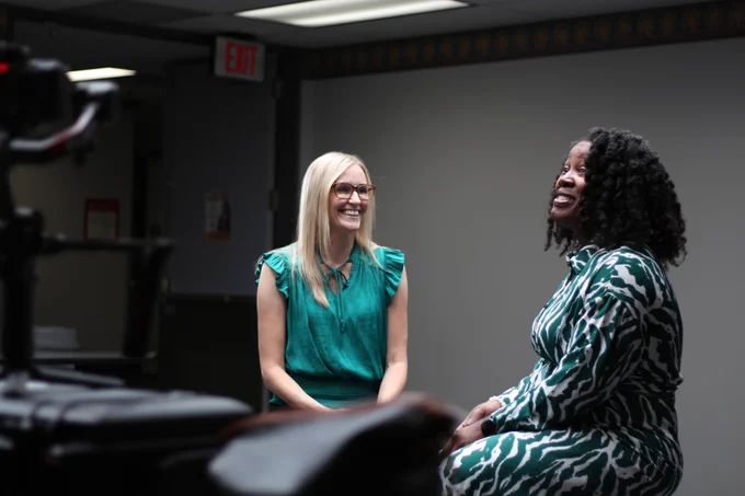 Dr. Meredith Masel, left, smiles while seated across from Dr. Mandy Hill during a filmed interview, with a video camera visible in the foreground.