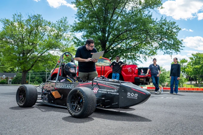 A driver in a racecar with someone next to them looking at a laptop, with three people in the background looking on