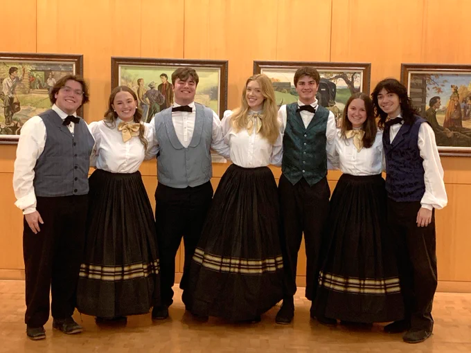 Group of six people dressed in traditional folk costumes standing arm in arm in front of framed paintings on a wooden wall.