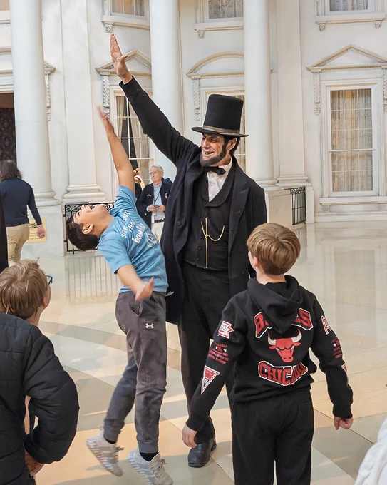 A man dressed as Abraham Lincoln gives a high-five to an excited boy jumping up indoors with two other children watching nearby.