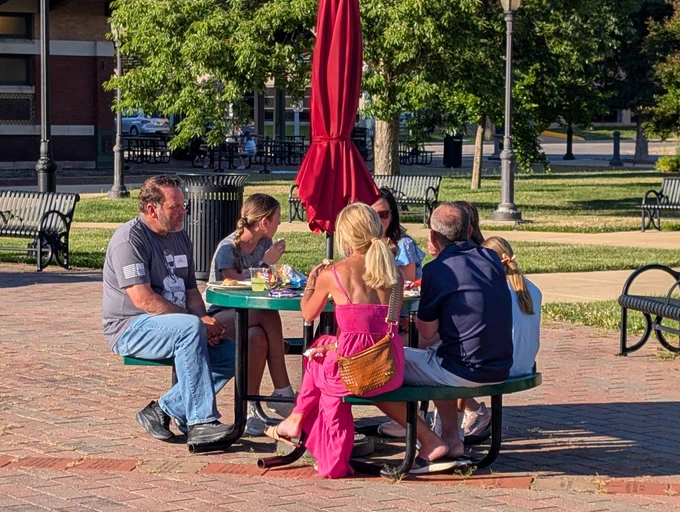 Group of six people sitting around a green picnic table with a closed red umbrella in a sunny park setting.