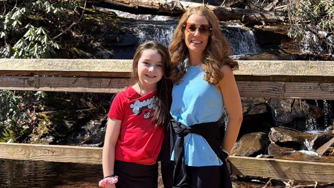 Jennifer Capouya and her daughter smile while standing on a wooden bridge in front of a waterfall surrounded by trees and rocks on a sunny day.